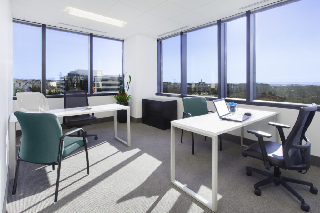 002 DO - Metro Offices A bright office with two desks, chairs, laptops, and a potted plant. Large windows provide natural light and a view of city buildings.