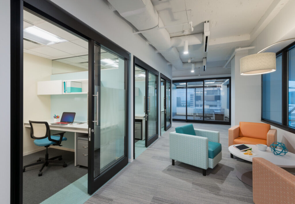 micro - Metro Offices Modern office space with glass-walled private offices, ergonomic chairs, a communal seating area with armchairs, and a small table. The large windows provide ample natural light, making it ideal for those working in shared spaces.