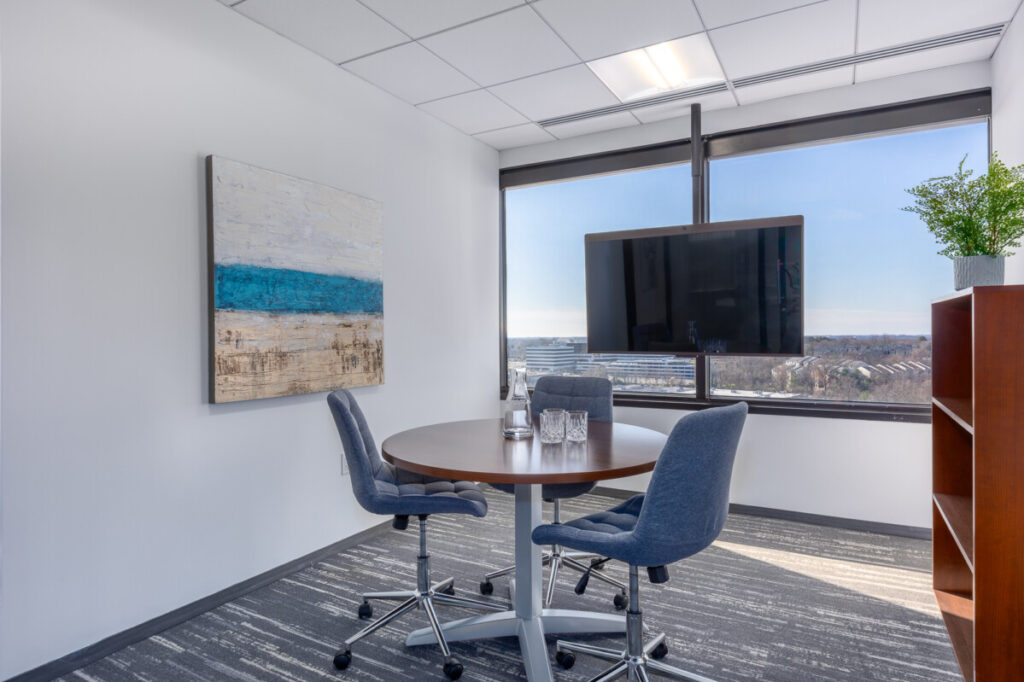 MetroOffices Tysons 38 - Metro Offices A small office with a round table, three chairs, a mounted TV, a framed painting on the wall, and a bookshelf with a potted plant. It's the perfect setup for those used to working in shared environments, enhanced by a window offering an impressive city view.