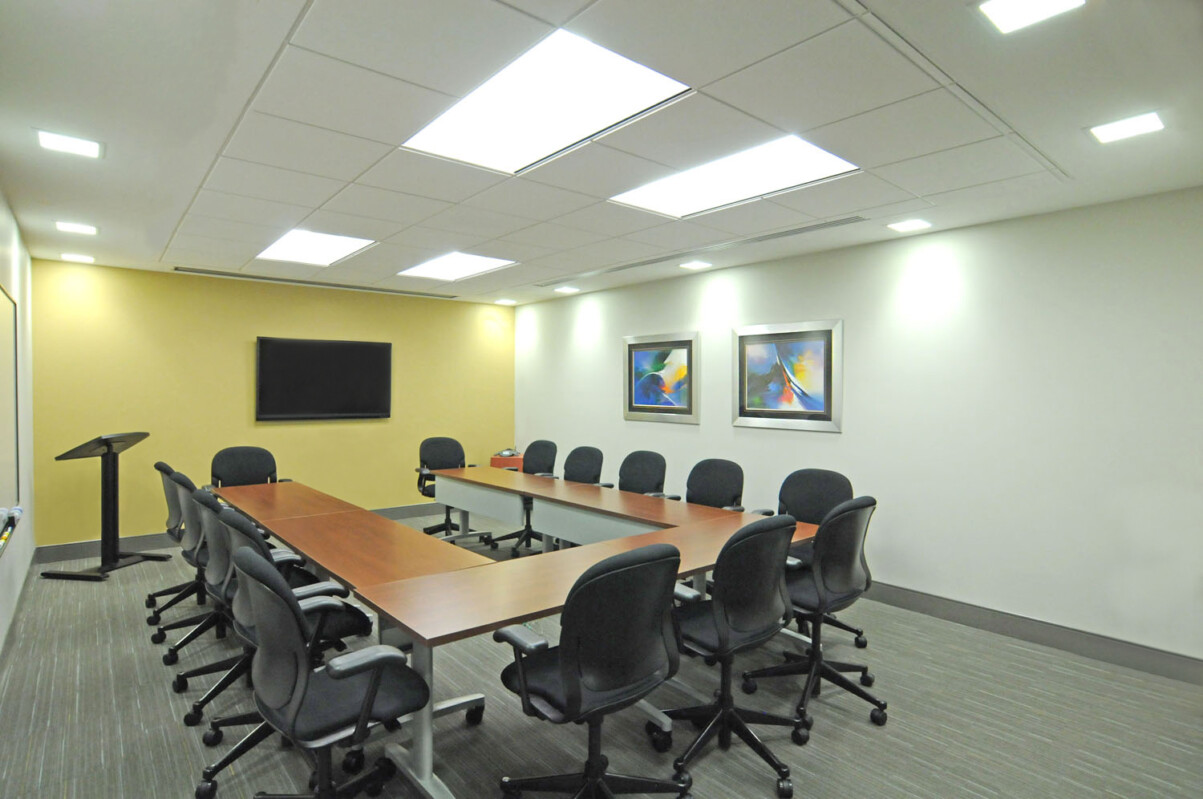 A conference room with a long rectangular table, several black office chairs, a podium, a wall-mounted TV, and two pieces of framed artwork on the wall.