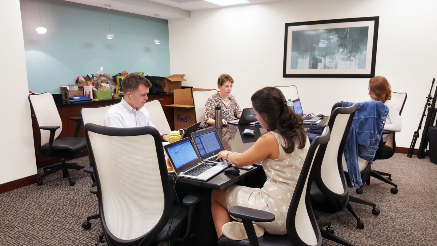Four people work on laptops around a conference table in an office environment. The room has a framed picture on the wall, a light blue accent wall, and several boxes in the corner.
