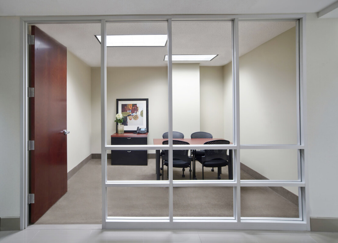 A modern office meeting room with a glass wall, a wooden door, a framed picture on the wall, a desk with flowers and a phone, and five chairs around a conference table.