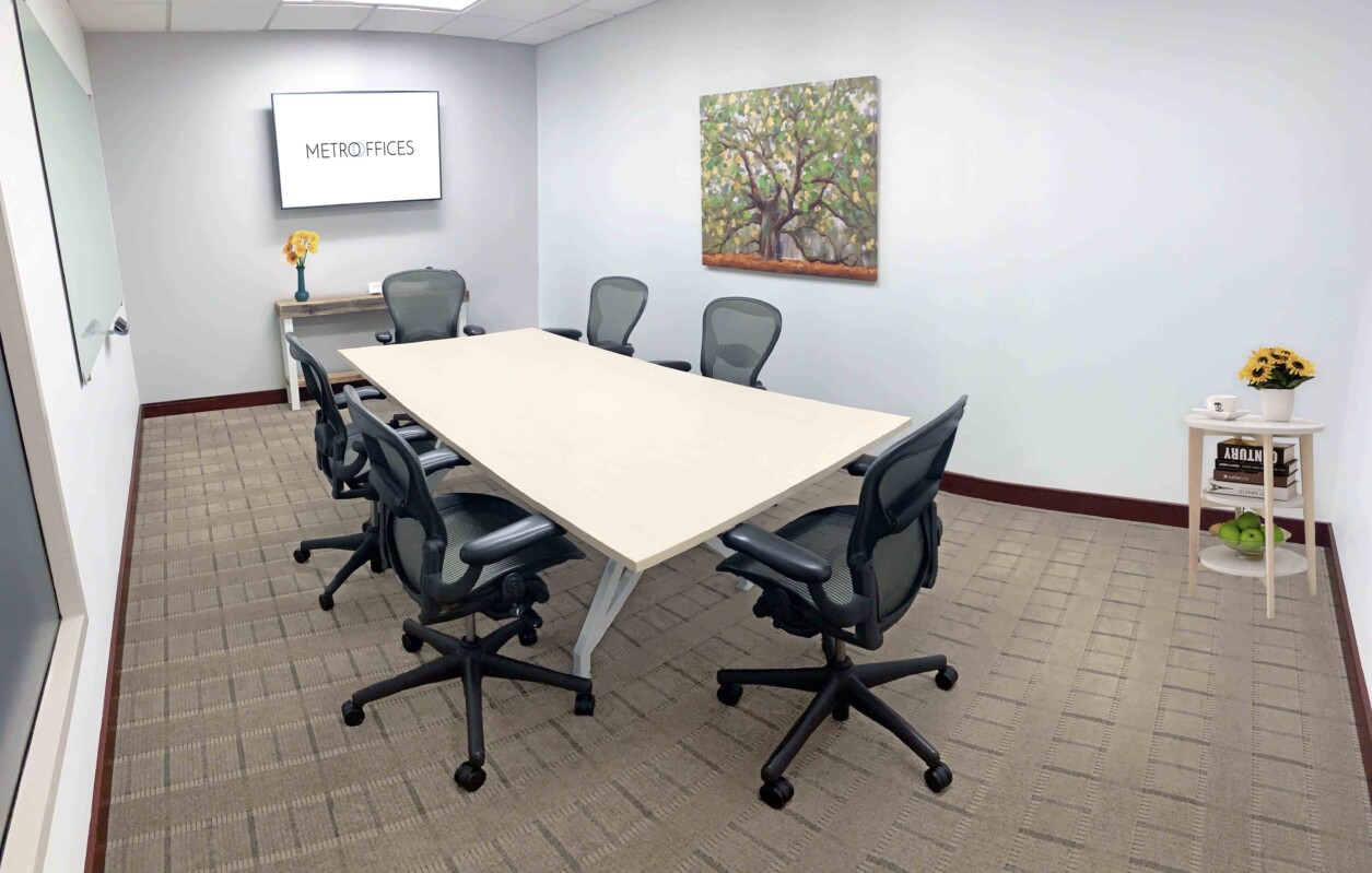 A small conference room with a rectangular table, six office chairs, a wall-mounted screen showing "Metro Offices," a painting, and a side table with sunflowers and a green fruit basket.