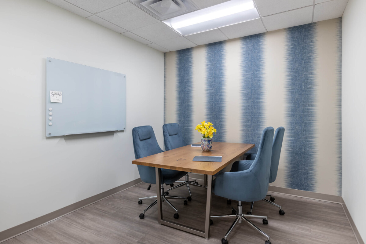 Small meeting room with a wooden table, four blue upholstered chairs, a whiteboard on the left wall, and blue striped accent wallpaper on the right. A vase with yellow flowers is on the table.