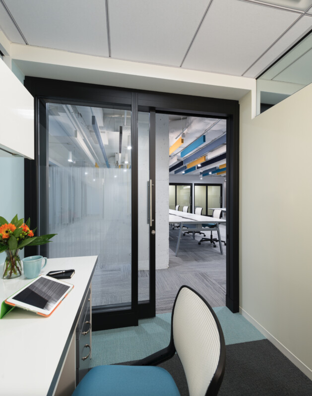 An office cubicle with a desk, chair, tablet, and flowers. A large glass sliding door separates the cubicle from a conference room with chairs and a long table.