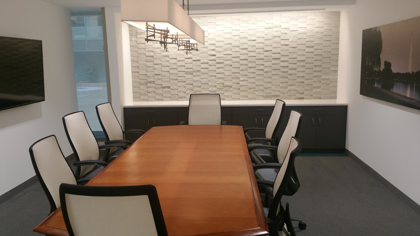 A conference room with a wooden table, six chairs, a wall-mounted light fixture, a textured wall, and a window. A television is mounted on one wall, and a piece of framed art is on another.