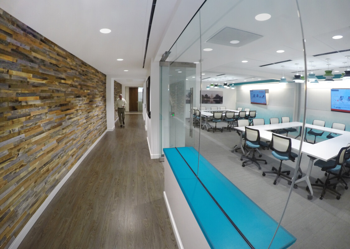 A modern office hallway with a wooden accent wall leads to a glass-partitioned conference room with chairs and large screens. A person walks down the hallway.