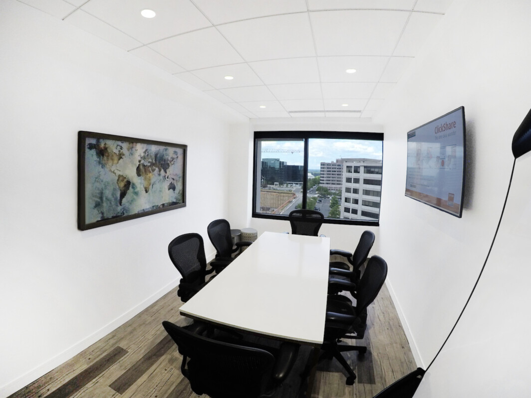 Modern conference room with a rectangular table, black office chairs, a large window showcasing an urban view, wall art, and two mounted screens.